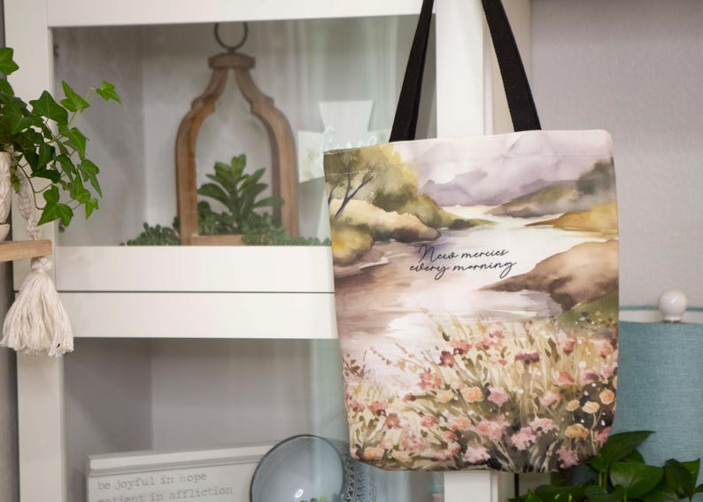 Puede incluir: Una bolsa de tela blanca con un dise&ntilde;o de paisaje de acuarela. La bolsa tiene correas negras y presenta el texto "New mercies every morning".