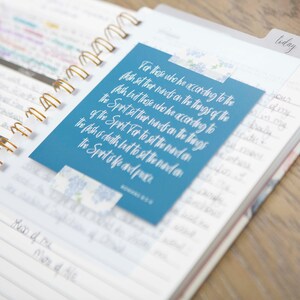 May include: A blue planner with a gold spiral binding. A blue card with white text is attached to the planner. The text on the card reads: "For those who he according to the flesh set their hands on the things of the flesh but those who he according to the Spirit set their hands on the things of the Spirit For to set the mind on the flesh is death but to set the mind on the Spirit of life and peace." Romans 8:6
