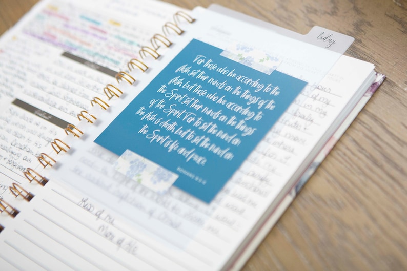 May include: A blue planner with a gold spiral binding. A blue card with white text is attached to the planner. The text on the card reads: "For those who he according to the flesh set their hands on the things of the flesh but those who he according to the Spirit set their hands on the things of the Spirit For to set the mind on the flesh is death but to set the mind on the Spirit of life and peace." Romans 8:6