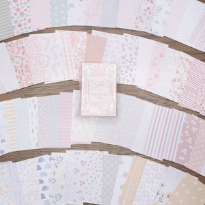 May include: A collection of patterned paper sheets in various designs and colors, including floral, striped, and dotted patterns. The color palette is soft, featuring pinks, whites, and pastel hues. A small card with text is centered among the sheets.