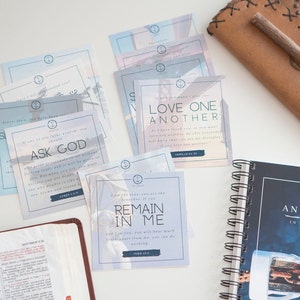 May include: A set of inspirational cards with bible verses and a blue and white watercolor background. The cards feature quotes like "Ask God", "Love One Another", and "Remain in Me". The cards are arranged on a white surface with a brown leather journal and a bible.