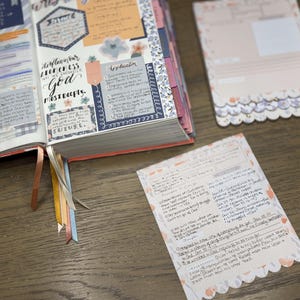 May include: An open journal with colorful decorative elements and handwritten notes. The pages feature floral accents, stickers, and ribbons. Two notecards with scalloped edges and handwritten text are also visible.