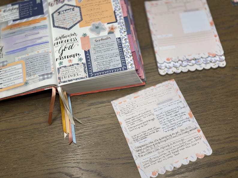 May include: An open journal with colorful decorative elements and handwritten notes. The pages feature floral accents, stickers, and ribbons. Two notecards with scalloped edges and handwritten text are also visible.