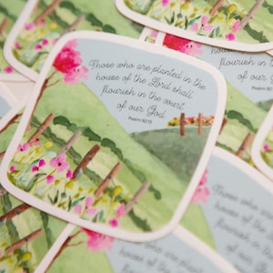 May include: A stack of green and white cards with a watercolor illustration of a field with pink flowers and a blue sky. The text on the card reads "Those who are planted in the house of the Lord shall flourish in the court of our God. Psalm 92:13."