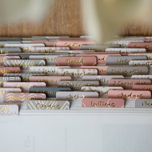 May include: A close-up of a white binder with gold-colored tabs labeled with the names of books in the Bible. The tabs are in a variety of patterns, including geometric and floral designs.