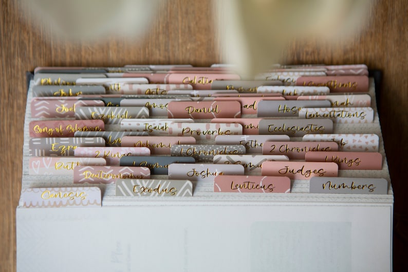 May include: A close-up of a white binder with gold-colored tabs labeled with the names of books in the Bible. The tabs are in a variety of patterns, including geometric and floral designs.