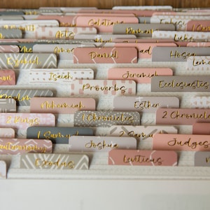 May include: A close-up of a set of pink and gray index cards with gold lettering. The cards are labeled with the names of books in the Bible, including Genesis, Exodus, Leviticus, Numbers, Deuteronomy, Joshua, Judges, Ruth, 1 Samuel, 2 Samuel, 1 Kings, 2 Kings, 1 Chronicles, 2 Chronicles, Ezra, Nehemiah, Esther, Job, Psalms, Proverbs, Ecclesiastes, Song of Solomon, Isaiah, Jeremiah, Lamentations, Ezekiel, Daniel, Hosea, Joel, Amos, Obadiah, Jonah, Micah, Nahum, Habakkuk, Zephaniah, Haggai, Zechariah, Malachi, Matthew, Mark, Luke, John, Acts, Romans, 1 Corinthians, 2 Corinthians, Galatians, Ephesians, Philippians, Colossians, 1 Thessalonians, 2 Thessalonians, 1 Timothy, 2 Timothy, Titus, Philemon, Hebrews, James, 1 Peter, 2 Peter, 1 John, 2 John, 3 John, Jude, and Revelation.