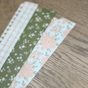 May include: Four decorative paper bookmarks with floral and geometric patterns. The patterns include a peach and white plaid, small white flowers on a green background, and peach flowers on a light blue background.