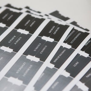 May include: A set of black and white book divider tabs with the names of books of the Bible printed on them. The tabs are designed to be used in a Bible to help organize and find specific books.