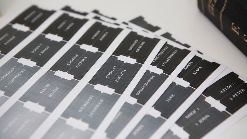 May include: A set of black and white book divider tabs with the names of books of the Bible printed on them. The tabs are designed to be used in a Bible to help organize and find specific books.