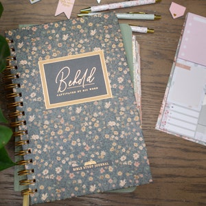 May include: A floral-patterned Bible study journal with the word "Behold" on the cover, along with coordinating pens and sticky notes. The journal has a spiral binding and a gold-colored ribbon bookmark. The background is a wooden surface.