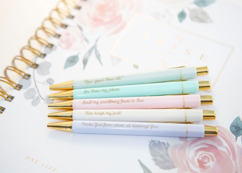 May include: Set of six pastel-colored pens with gold accents. Each pen has a different inspirational quote in gold lettering. The quotes are: "How great Thou art", "Be Thou my vision", "Bind my wandering heart to Thee", "Then sings my soul", "Praise God from whom all blessings flow".