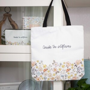 May include: White canvas tote bag with black handles and a floral print border. The bag has the text "Consider the wildflowers" printed in black.
