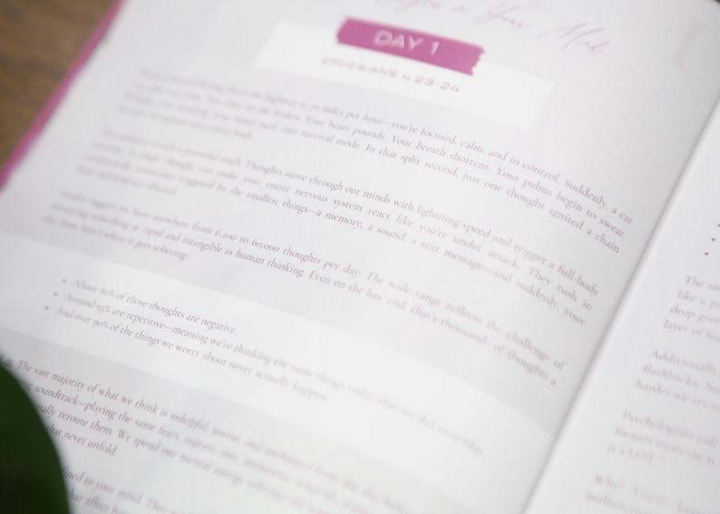 May include: Open book with text on white pages. The top of the page has a pink banner that says "DAY 1". The text is in a light gray color. The book is open to a page with text about thoughts and the mind.