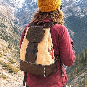 May include: Tan and brown leather backpack with a deer emblem. The backpack has a flap closure and a stitched design. The person is wearing a mustard yellow knit cap and a maroon jacket, standing in front of a mountain range.