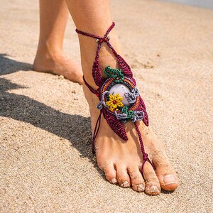 May include: A macrame barefoot sandal in burgundy, green, yellow, and purple. The sandal has a central circular design with a light-coloured stone and floral accents. Cords wrap around the foot. The image is taken on a sandy beach.