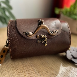 Pode incluir: Uma bolsa cilíndrica de couro marrom com um fecho de latão decorativo e uma alça. A bolsa tem um estilo vintage e é feita de couro de alta qualidade.