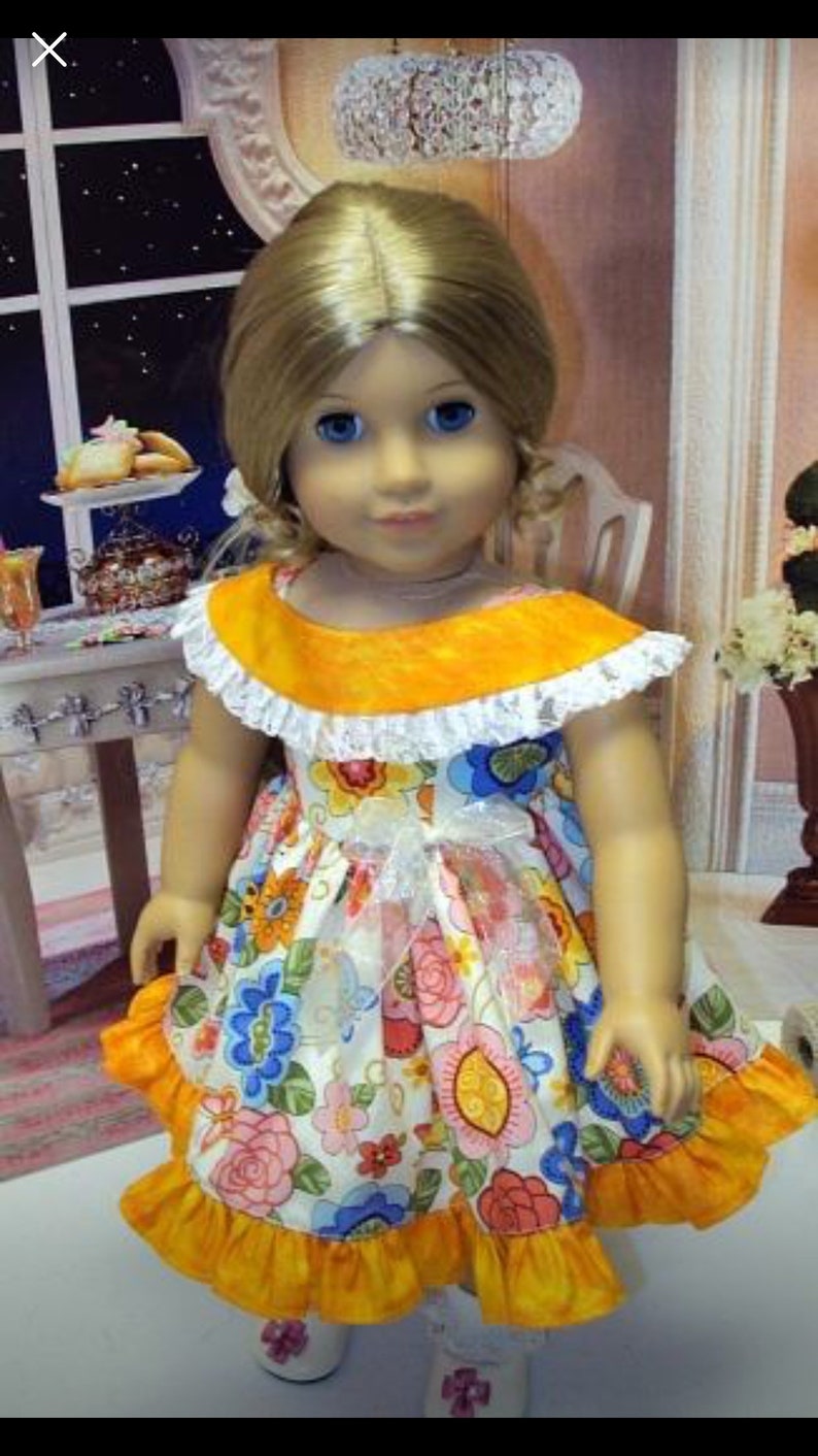 May include: A doll wearing a floral print dress with a yellow collar and ruffled hem. The dress features a white bow and is paired with white shoes. The doll has blonde hair and blue eyes.