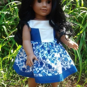 May include: A doll with long black hair wearing a blue and white floral dress and white shoes. The dress has a white bodice with blue accents and a blue hem. The doll is standing in a grassy area.