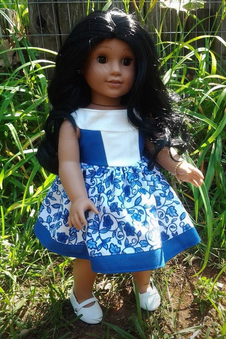 May include: A doll with long black hair wearing a blue and white floral dress and white shoes. The dress has a white bodice with blue accents and a blue hem. The doll is standing in a grassy area.
