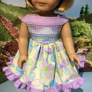 May include: A doll wearing a handmade dress with a floral pattern. The dress features a light green background with large purple, blue, and green flowers. The bodice is a mix of purple polka dots and checkered patterns. The dress has a ruffled hem.