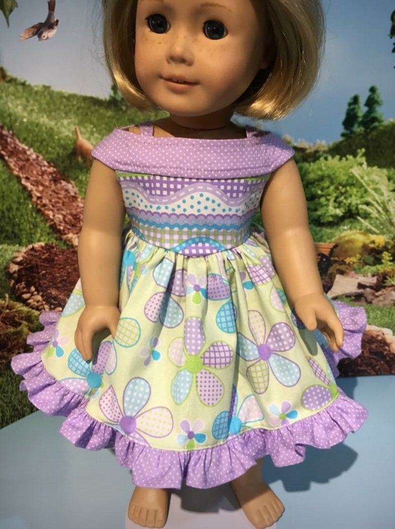 May include: A doll wearing a handmade dress with a floral pattern. The dress features a light green background with large purple, blue, and green flowers. The bodice is a mix of purple polka dots and checkered patterns. The dress has a ruffled hem.