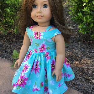 May include: A doll with brown hair and blue eyes wearing a turquoise dress with a floral pattern of pink and white flowers. The dress has a ruffled hem and straps. The doll is standing outside in front of green foliage.