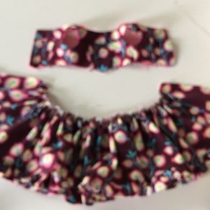 May include: A two-piece set of children's clothing. The top is a bandeau style, and the bottom is a ruffled skirt. Both pieces are made of a maroon fabric with a floral pattern in pink, yellow, and teal.
