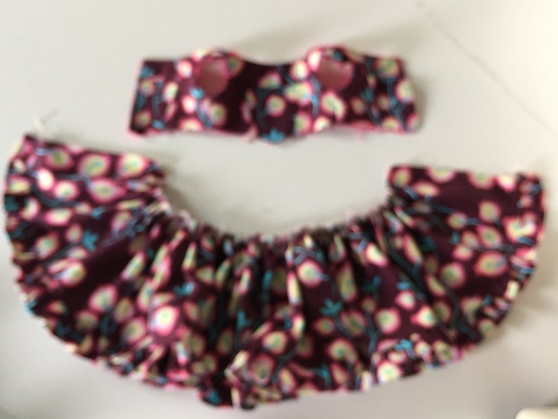 May include: A two-piece set of children's clothing. The top is a bandeau style, and the bottom is a ruffled skirt. Both pieces are made of a maroon fabric with a floral pattern in pink, yellow, and teal.