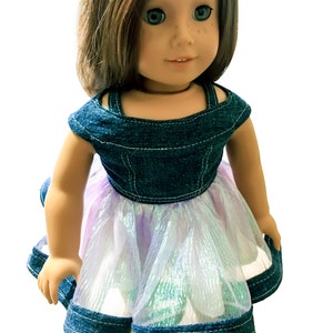 May include: A doll wearing a denim bodice dress with a layered, iridescent skirt. The dress features a square neckline and is trimmed with denim. The doll has brown hair, blue eyes, and freckles.