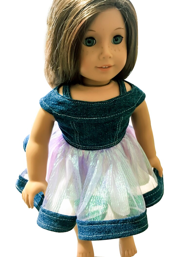 May include: A doll wearing a denim bodice dress with a layered, iridescent skirt. The dress features a square neckline and is trimmed with denim. The doll has brown hair, blue eyes, and freckles.