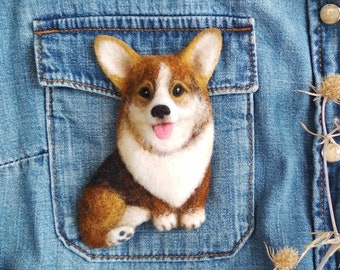 May include: A felt brooch of a brown and white corgi dog with a pink tongue sticking out. The dog is sitting with its front paws crossed and is looking directly at the camera.