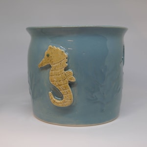 May include: A light blue ceramic jar with a yellow seahorse design. The jar has a textured surface and is perfect for storing small items.
