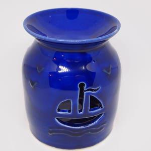 May include: A blue ceramic oil burner with a cutout design of a sailboat on the front. The burner has a wide, flat top for holding a tea light.