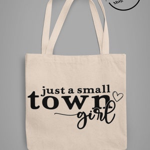 May include: A beige tote bag with black handles and the text "just a small town girl" with a small heart graphic.