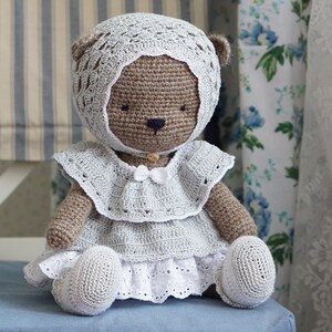 May include: A crocheted brown teddy bear wearing a white crocheted dress and hat. The bear is sitting on a blue chair.