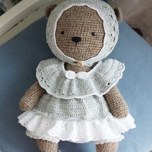 May include: A crocheted brown teddy bear wearing a light blue dress with white lace trim and a matching bonnet. The bear has black eyes and a button nose.