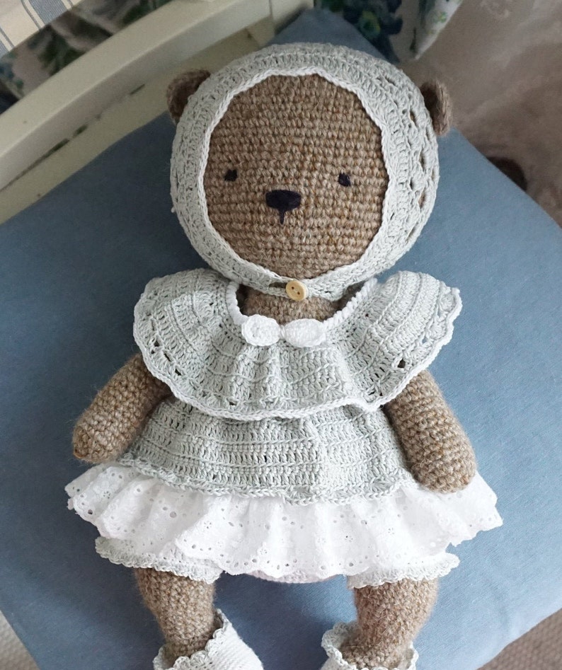 May include: A crocheted brown teddy bear wearing a light blue dress with white lace trim and a matching bonnet. The bear has black eyes and a button nose.