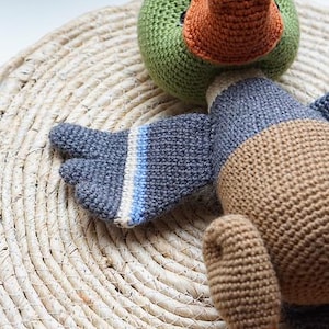 Op de afbeelding: Een gehaakt eendenspeelgoed met een groene kop en een oranje snavel. De vleugel is blauw en grijs met witte en blauwe strepen. Het speelgoed ligt op een geweven strooien mat. De tekst "Mariia Ermolova Polushkabunny" staat linksboven.