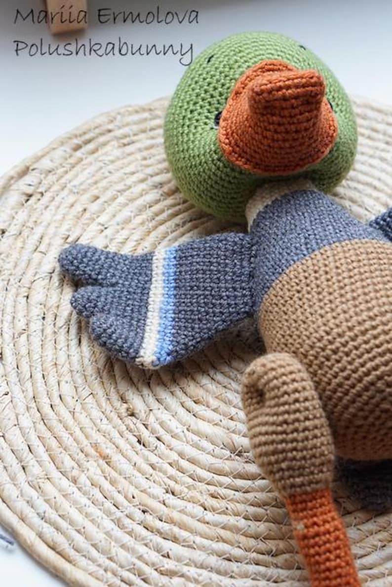 Op de afbeelding: Een gehaakt eendenspeelgoed met een groene kop en een oranje snavel. De vleugel is blauw en grijs met witte en blauwe strepen. Het speelgoed ligt op een geweven strooien mat. De tekst "Mariia Ermolova Polushkabunny" staat linksboven.