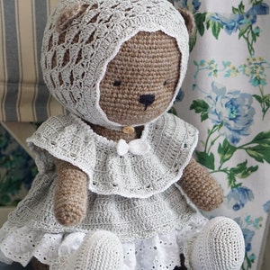 May include: A crocheted teddy bear wearing a white crocheted dress and bonnet. The bear has brown fur and black eyes.