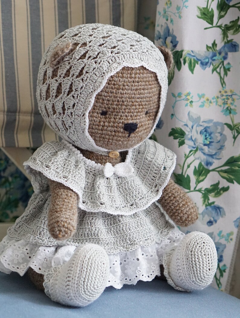 May include: A crocheted teddy bear wearing a white crocheted dress and bonnet. The bear has brown fur and black eyes.