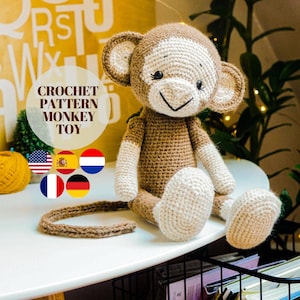 Op de afbeelding: Haakpatroon voor een bruin en wit aapje. Het patroon is beschikbaar in het Engels, Spaans, Nederlands, Frans, Duits en Italiaans. De aap zit op een tafel met een boek en een gele bol wol op de achtergrond.