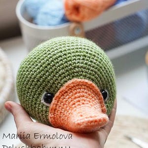 Op de afbeelding: Een handgemaakte, groene gehaakte eend met een oranje snavel en zwarte ogen. De eend wordt in een hand gehouden. De tekst "Mariia Ermolova Polushkabunny" staat op de afbeelding. Garen in blauwe en oranje tinten staat op de achtergrond.