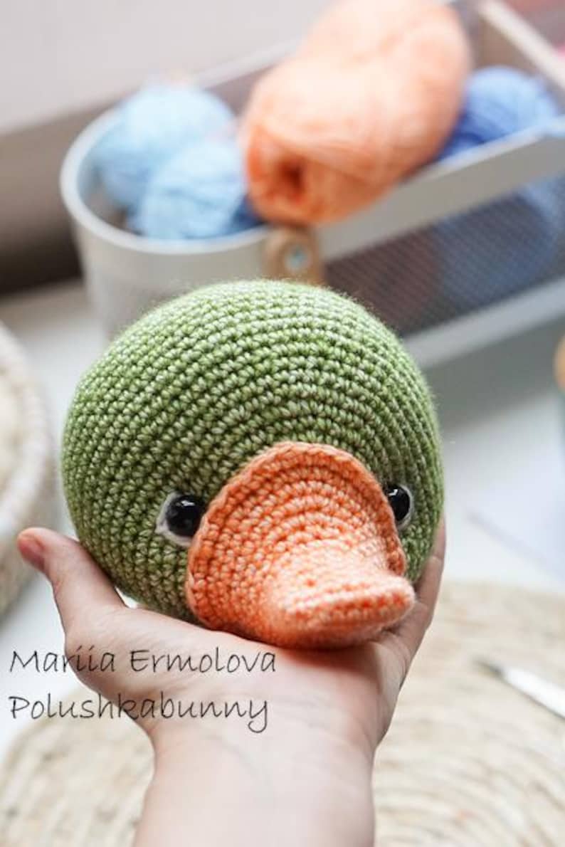 Op de afbeelding: Een handgemaakte, groene gehaakte eend met een oranje snavel en zwarte ogen. De eend wordt in een hand gehouden. De tekst "Mariia Ermolova Polushkabunny" staat op de afbeelding. Garen in blauwe en oranje tinten staat op de achtergrond.