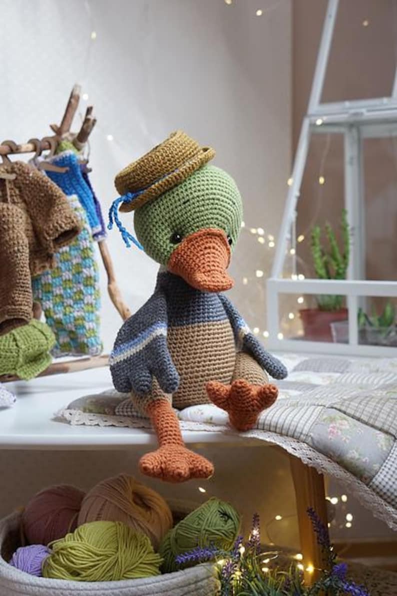 Op de afbeelding: Een handgemaakte gehaakte eendenpop met een groene kop, bruine hoed en oranje snavel en voeten. De eend heeft een blauw en bruin lichaam met blauwe strepen. De pop zit op een wit oppervlak met garen en andere haakartikelen.