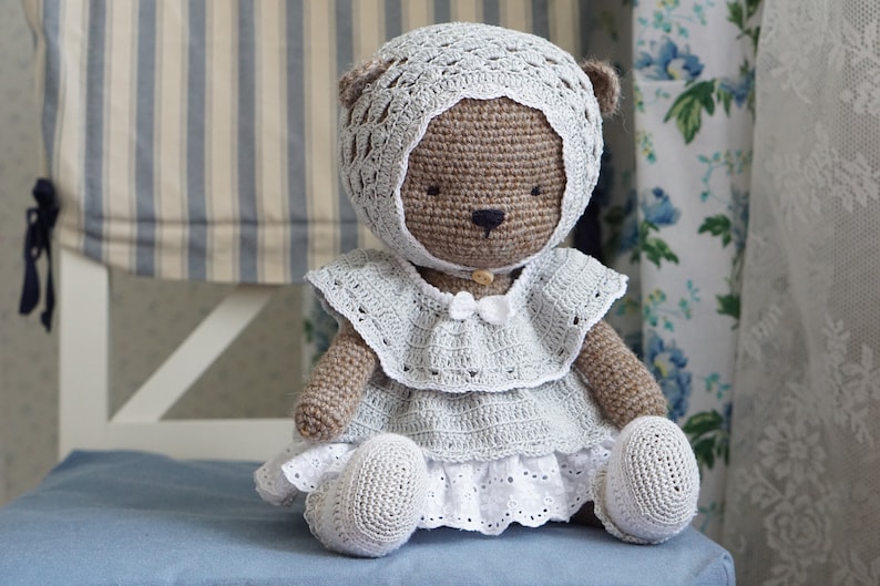 May include: A crocheted brown teddy bear wearing a white crocheted dress and hat. The bear is sitting on a blue chair.