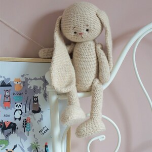 May include: A crocheted beige bunny rabbit toy with long ears, sitting on a white metal bed frame. The bunny has black eyes and a pink nose.