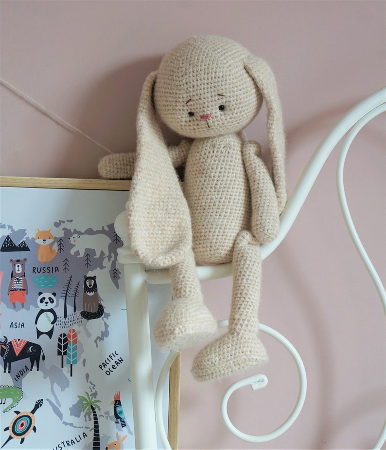 May include: A crocheted beige bunny rabbit toy with long ears, sitting on a white metal bed frame. The bunny has black eyes and a pink nose.