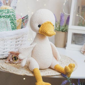 May include: A handmade crochet duck toy featuring a cream-colored body, a bright yellow beak, and matching yellow webbed feet. The stuffed animal has small black bead eyes and is crafted with a textured yarn stitch throughout its entire form.